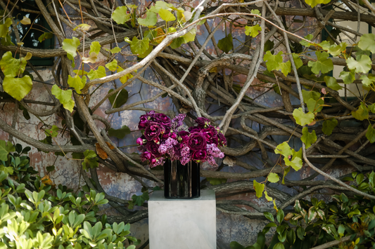 A flower arrangement to celebrate Mother's Day, featuring purple lilacs and Garden Roses from Rose Story Farm. Set in a background of vines and a patina wall.