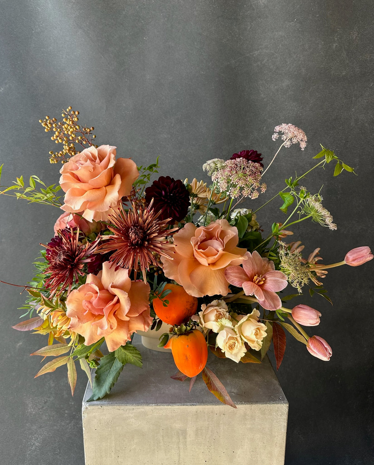 An autumnal floral arrangement featuring peach roses, dark burgundy chrysanthemums, pink tulips, and orange fruits, displayed on a concrete pedestal against a dark gray textured background.