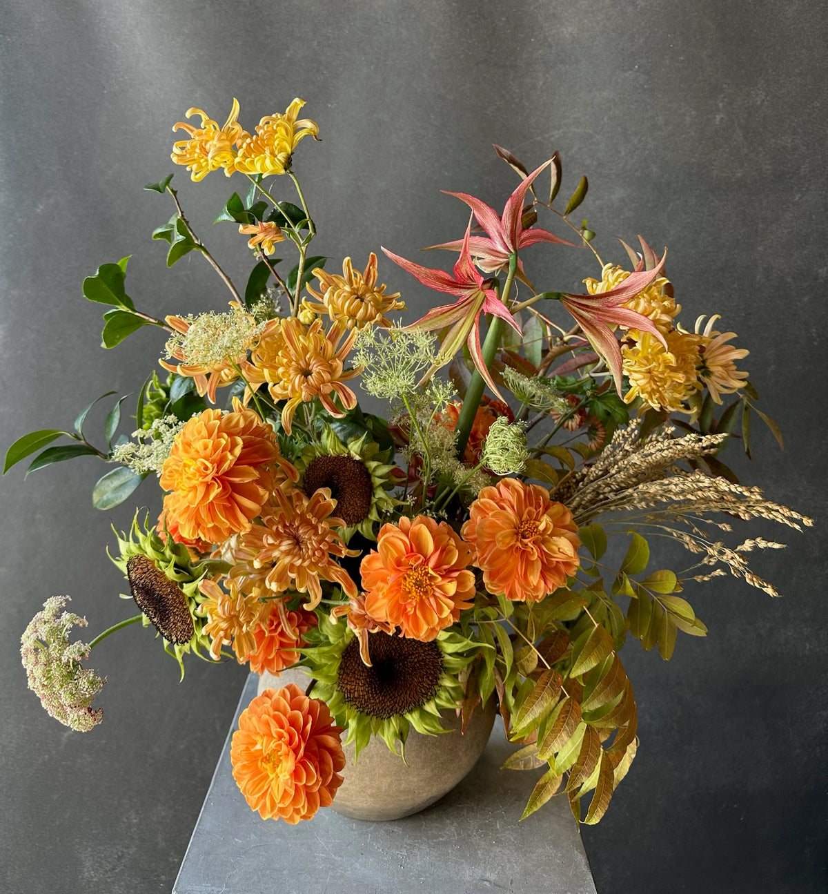 A vibrant autumnal floral arrangement in a round beige ceramic pot, featuring orange dahlias, yellow flowers, green foliage, and dried grasses, set against a dark gray textured background.