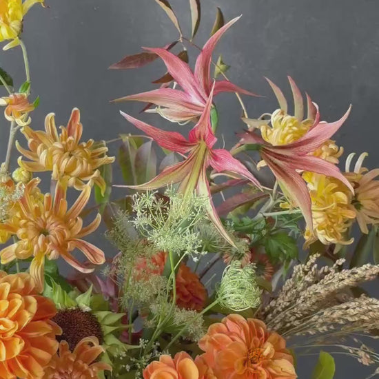 Tall autumn statement floral arrangement featuring golden chrysanthemums, orange dahlias, sculptural pink blooms, sunflowers, seasonal grasses, and autumn foliage in a ceramic vessel.