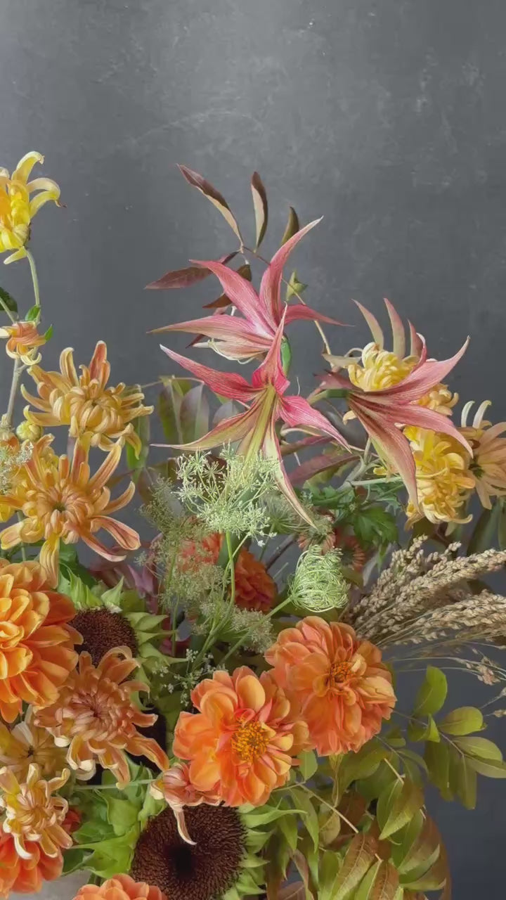 Tall autumn statement floral arrangement featuring golden chrysanthemums, orange dahlias, sculptural pink blooms, sunflowers, seasonal grasses, and autumn foliage in a ceramic vessel.