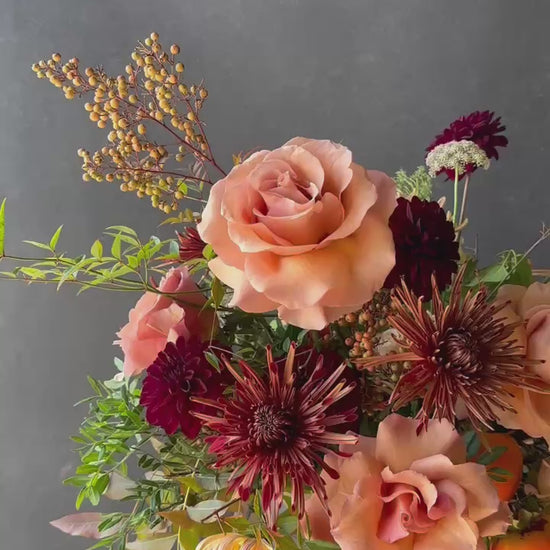 Thanksgiving floral centerpiece featuring peach garden roses, burgundy dahlias, blush blooms, seasonal persimmons, and autumn foliage in a low arrangement.