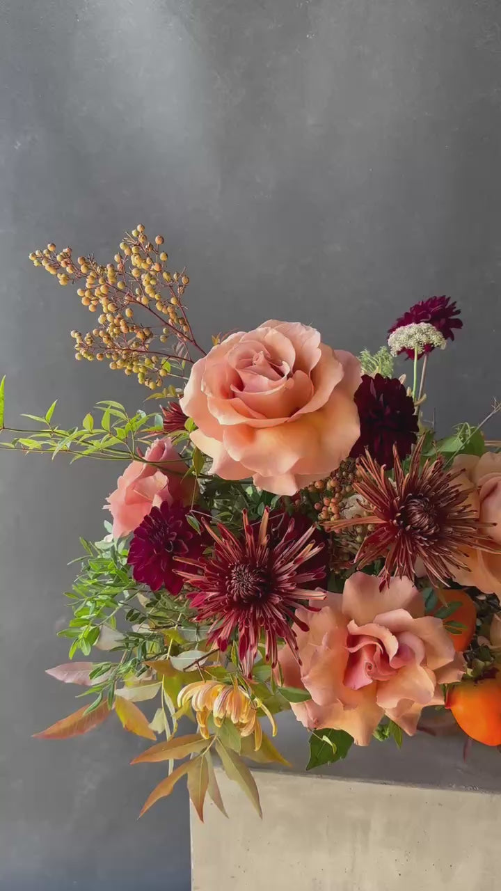 Thanksgiving floral centerpiece featuring peach garden roses, burgundy dahlias, blush blooms, seasonal persimmons, and autumn foliage in a low arrangement.